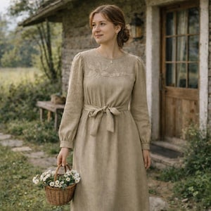 May include: A long-sleeved, beige dress with a tie-waist and floral embroidery on the bodice. The dress is paired with a small, woven basket filled with white flowers. The dress is made of a textured fabric, and the overall style is rustic and elegant.