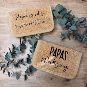 Könnte beinhalten: Zwei rechteckige Korkbeutel mit schwarzem Text. Einer lautet "Papa wird's schon richten!" und der andere "PAPAS Werkzeug". Die Beutel liegen auf einer Holzoberfläche mit Eukalyptusblättern.