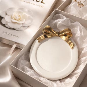 May include: A white ceramic plate with a gold bow detail, presented in a white box with tissue paper. A book titled "CHANEL COLLECTIONS AND CREATIONS" is visible, along with a white flower. The overall aesthetic is elegant and luxurious.
