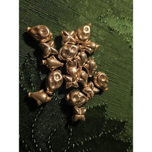 May include: A cluster of gold-colored charms in various shapes, including butterflies, faces, and abstract forms. The charms are arranged on a dark green, textured surface, creating a decorative display.