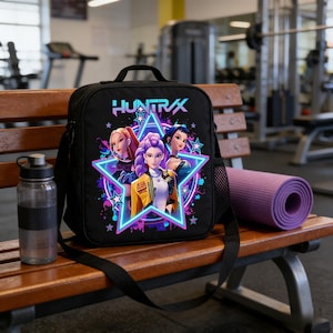 May include: Black lunch bag with a shoulder strap, featuring a colorful graphic of three anime-style women inside a star with the text "HUNTRAX". A clear water bottle and a rolled-up purple yoga mat are next to the bag on a wooden bench.