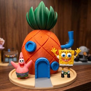 Może przedstawiać: Model 3D domu z ananasa SpongeBoba, z figurkami Patryka Rozgwiazdy i SpongeBoba. Dom jest pomarańczowy z zielonym dachem, niebieskimi akcentami i niebieską rurą. Figurki stoją na beżowej podstawie.