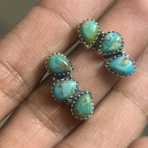 May include: A pair of turquoise earrings. Each earring features three teardrop-shaped turquoise stones set in a silver-colored metal. The turquoise stones have a mottled appearance with shades of blue and green.