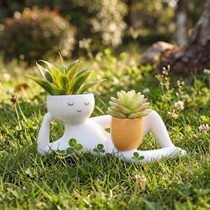 May include: A white ceramic planter shaped like a reclining figure with a closed-eye face, holding a green succulent in its head and a smaller, separate orange pot containing a light green succulent resting on its leg.