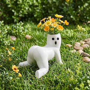 May include: A white ceramic planter shaped like a figure on all fours with a rounded posterior and a head featuring two black circular eyes and a small black circular mouth, filled with a bouquet of small yellow and orange flowers.