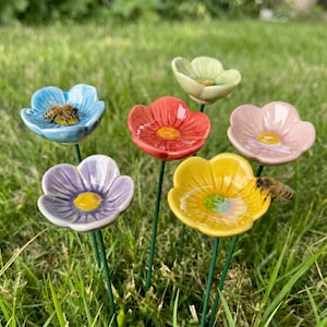 May include: A collection of colorful ceramic flower-shaped bee waterers. Each flower has a different color, including blue, red, pink, yellow, purple, and green, with a yellow center. Bees are present on two of the flowers. The flowers are on green stems.
