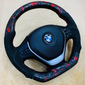 May include: A black and red steering wheel with the BMW logo in the center. The wheel has a carbon fiber design with red accents and black suede-like grips. Red stitching is visible.