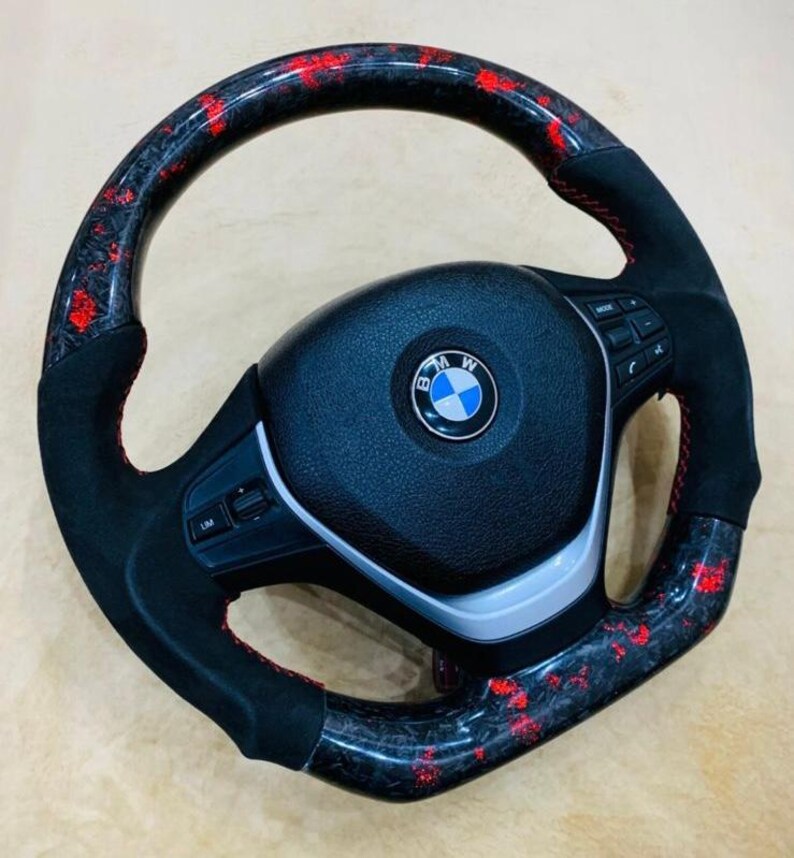 May include: A black and red steering wheel with the BMW logo in the center. The wheel has a carbon fiber design with red accents and black suede-like grips. Red stitching is visible.