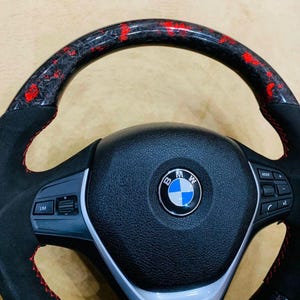 May include: A black and red BMW steering wheel with a carbon fiber and red accent top. The wheel has a black leather center with the BMW logo and red stitching. The wheel has buttons for mode and other functions.