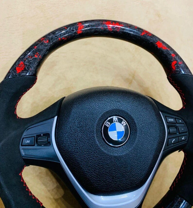 May include: A black and red BMW steering wheel with a carbon fiber and red accent top. The wheel has a black leather center with the BMW logo and red stitching. The wheel has buttons for mode and other functions.