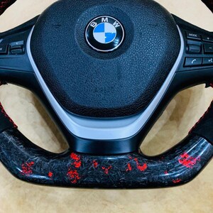 May include: Close-up of a black and red steering wheel. The wheel features a carbon fiber design with red accents. The center displays the BMW logo. The wheel has buttons and controls on the sides.