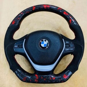 May include: A black and red carbon fiber steering wheel with a BMW logo. The wheel has a black suede grip with red stitching. The carbon fiber sections have a red speckled pattern.