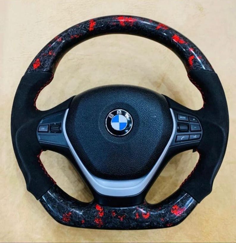 May include: A black and red carbon fiber steering wheel with a BMW logo. The wheel has a black suede grip with red stitching. The carbon fiber sections have a red speckled pattern.