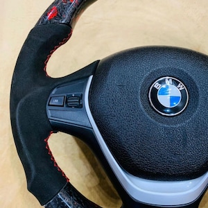 May include: A close-up of a black and red carbon fiber steering wheel. The wheel has a black suede grip with red stitching. The center features the BMW logo.