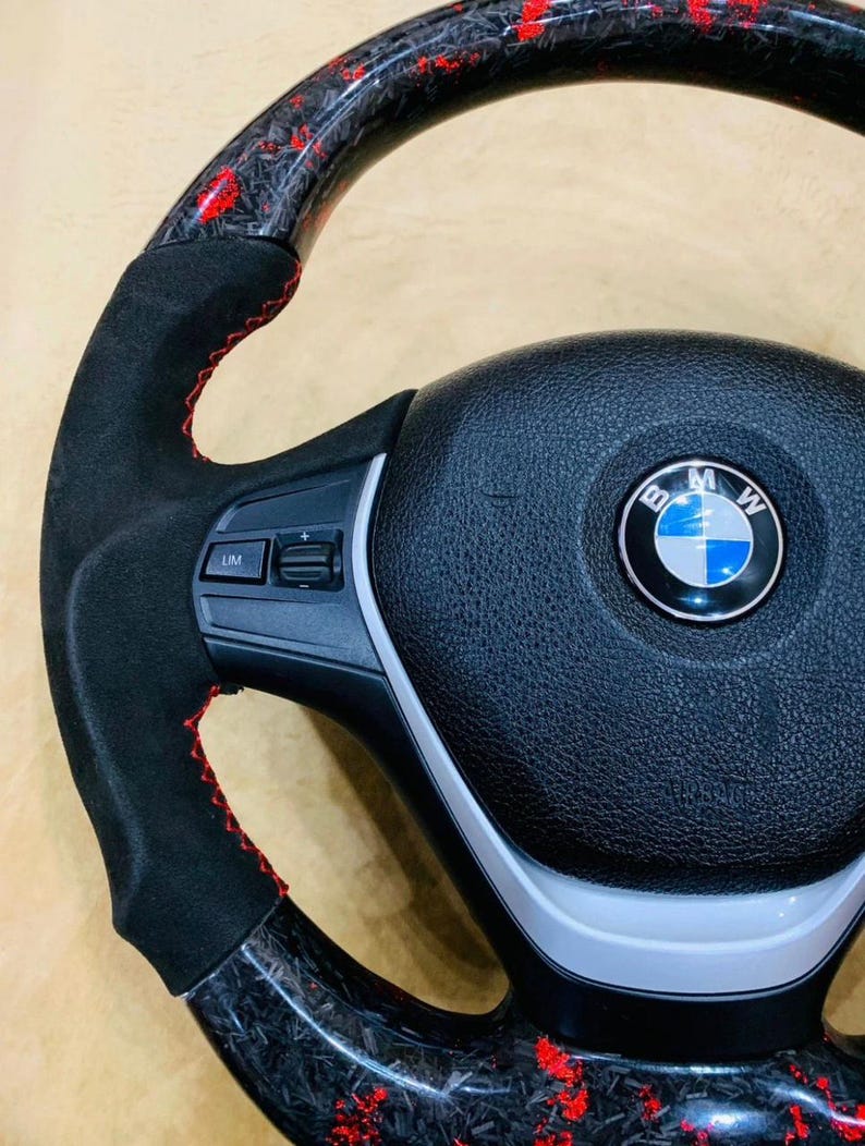 May include: A close-up of a black and red carbon fiber steering wheel. The wheel has a black suede grip with red stitching. The center features the BMW logo.