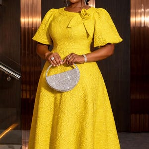 May include: A vibrant yellow dress with a textured design, featuring short, flared sleeves and a round neckline. A sparkling, half-moon shaped clutch is held in front. A floral accent adorns the bodice. The dress is knee-length.