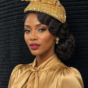May include: A woman wearing a gold-colored, sequined headpiece with a small, decorative gold ball on top. She is also wearing a gold satin blouse with a bow tie. The background is a black, textured surface.
