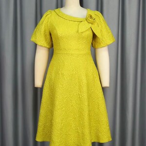 May include: A vibrant yellow dress with a floral texture, short sleeves, and a rounded neckline. The dress has a decorative rose detail on the shoulder and a fitted bodice that flows into an A-line skirt. The dress is displayed on a mannequin.