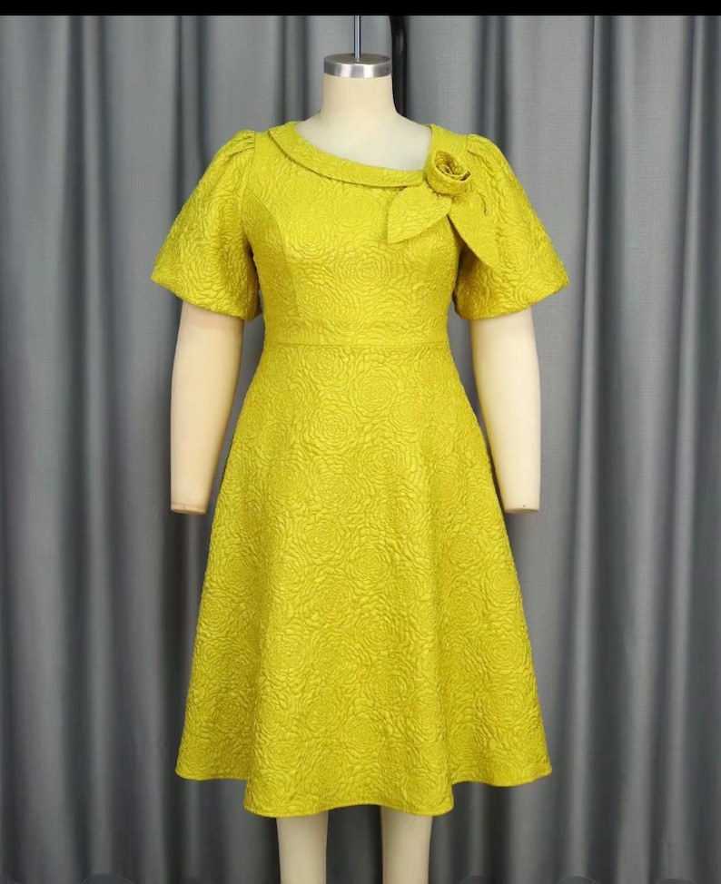 May include: A vibrant yellow dress with a floral texture, short sleeves, and a rounded neckline. The dress has a decorative rose detail on the shoulder and a fitted bodice that flows into an A-line skirt. The dress is displayed on a mannequin.