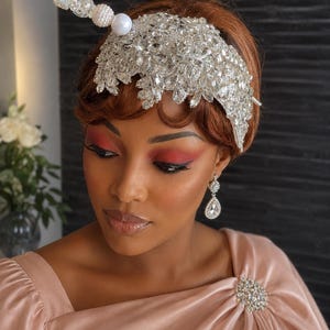 May include: A silver-toned headpiece adorned with clear crystals and a decorative stick featuring white and clear beads. The accessory is paired with silver-toned teardrop earrings and a light pink satin garment featuring a circular crystal brooch.