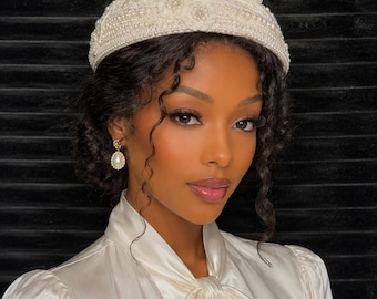 May include: Ivory-colored, beaded hat with a decorative beaded accent. The hat is adorned with pearls and small floral embellishments. The woman in the image is wearing a white satin blouse with a bow.