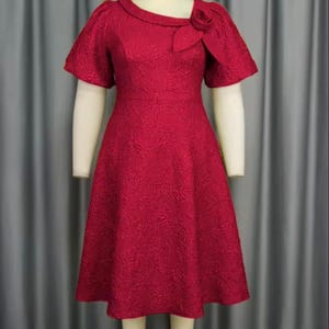 May include: A red knee-length dress with a textured floral pattern. The dress has short sleeves, a unique neckline, and a decorative rose detail on the shoulder. The dress is displayed on a mannequin.