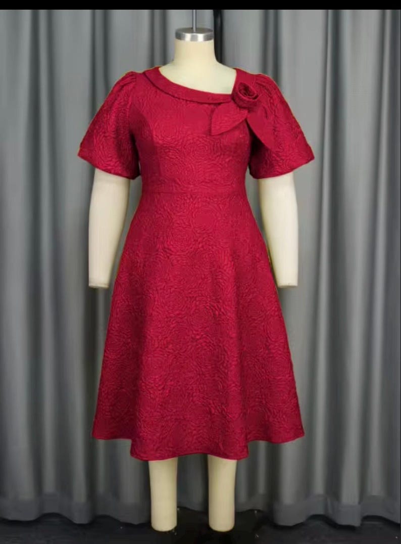 May include: A red knee-length dress with a textured floral pattern. The dress has short sleeves, a unique neckline, and a decorative rose detail on the shoulder. The dress is displayed on a mannequin.