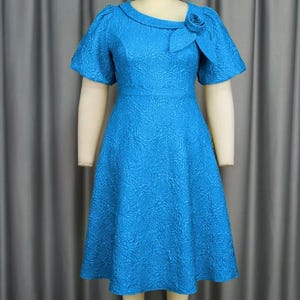 May include: A bright turquoise dress with a textured floral design. The dress has short sleeves, a fitted waist, and a flared skirt. A decorative rose detail is on the neckline. The dress is displayed on a mannequin.