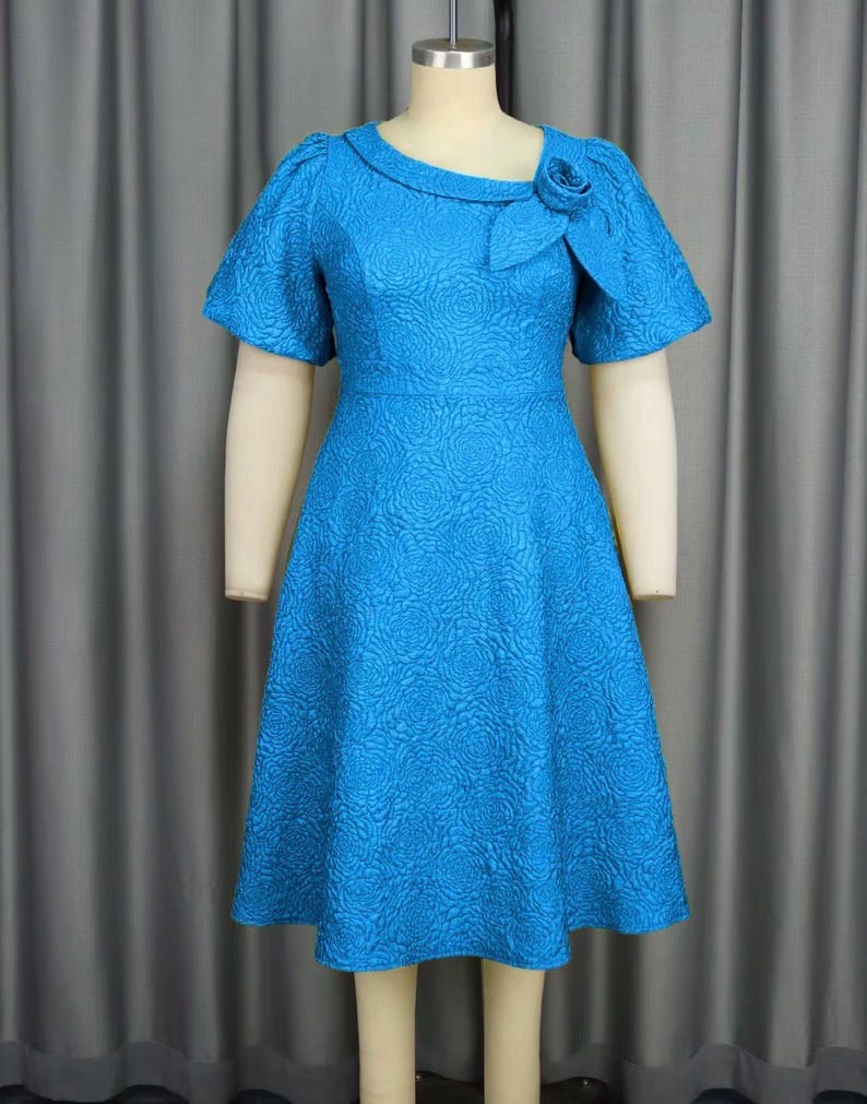May include: A bright turquoise dress with a textured floral design. The dress has short sleeves, a fitted waist, and a flared skirt. A decorative rose detail is on the neckline. The dress is displayed on a mannequin.