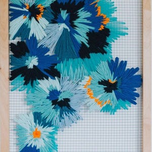 Op de afbeelding: Een ingelijst textielkunstwerk met abstracte bloemmotieven. De bloemen zijn gemaakt van garen in verschillende tinten blauw, turquoise en zwart, met oranje accenten. Het kunstwerk is geplaatst tegen een rasterachtergrond en ingelijst in natuurlijk hout.