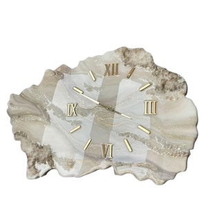 Puede incluir: Un reloj de pared decorativo con una forma orgánica única. La esfera del reloj presenta un patrón veteado en tonos blancos, beige y dorados. Los números romanos dorados marcan las horas, y las manecillas doradas indican la hora.