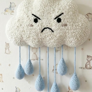 Może przedstawiać: Ręcznie robiona, biała szydełkowa dekoracja ścienna w kształcie chmury z wściekłą twarzą, czarnymi oczami i niebieskimi kroplami deszczu. Chmura zawieszona jest na drewnianym kołku i makramie. Tło to tapeta z ilustracjami zwierząt.