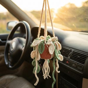 May include: A crocheted hanging plant in a car. The plant pot is a reddish-brown color, with green and off-white leaves. The plant hangs from the rearview mirror by three beige cords. The car interior is visible, with a steering wheel and dashboard.