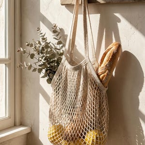 May include: A beige, open-weave shopping bag hangs from a wooden hook on a white wall. The bag contains a loaf of bread and several yellow fruits. Greenery is tucked into the bag's side. The scene is lit by natural light.
