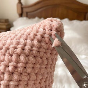May include: Close-up of a light pink, chunky knit pillow. A pair of silver scissors is cutting a small piece of the pillow's material. The pillow is on a white surface, with a wooden headboard in the background.