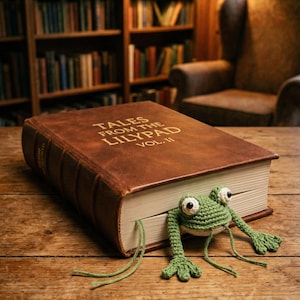 May include: A large, brown leather-bound book titled "TALES FROM THE LILYPAD VOL. II" with a green crocheted frog bookmark. The frog has large eyes and long legs. The book rests on a wooden table, with a bookshelf and armchair in the background.