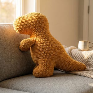 May include: A mustard-colored, crocheted dinosaur plush toy. The dinosaur is positioned on a gray sofa, with its head and arms resting on the sofa's arm. The plush has a textured surface and is near a window.