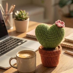 Pode incluir: Um cacto em forma de coração de crochê com uma flor rosa em um vaso laranja em uma mesa de madeira. Uma xícara de café e um laptop estão por perto. Uma planta suculenta em um vaso e cadernos também estão na mesa.