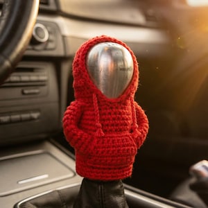 Peut inclure: Un sweat à capuche rouge en crochet recouvre le pommeau de levier de vitesses d'une voiture. La housse faite à la main a un capuchon avec des cordons et est faite d'un fil rouge texturé. Le levier de vitesses est à l'intérieur d'une voiture.
