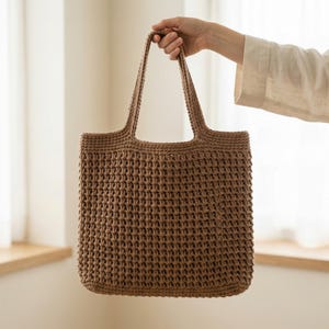 Könnte beinhalten: Eine braune Häkeltasche mit zwei Henkeln. Die Tasche wird von einer Hand gehalten. Die Tasche hat eine rechteckige Form und ein strukturiertes, gewebtes Aussehen. Die Tasche ist aus einem dicken, strapazierfähigen Garn gefertigt.