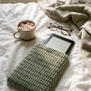 Puede incluir: Una funda de tableta de ganchillo verde salvia con una tableta dentro, sobre una cama blanca. Una taza de chocolate caliente con malvaviscos y un par de gafas están cerca. La escena evoca un ambiente acogedor y relajante.