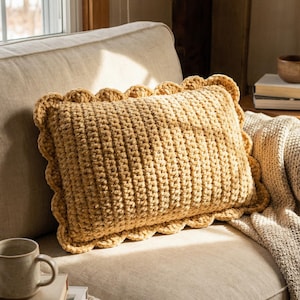 May include: A mustard-coloured crochet cushion with a scalloped edge sits on a beige sofa. The textured surface of the cushion is visible, and it is surrounded by a neutral-toned blanket. A mug and books are nearby.