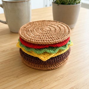 Puede incluir: Un conjunto de posavasos de ganchillo hechos a mano con forma de hamburguesa. La "hamburguesa" presenta capas marrones, amarillas, verdes y rojas, coronadas con un "pan" marrón. Los posavasos están sobre una superficie de madera clara.