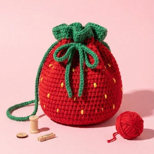 May include: A red crocheted strawberry-shaped drawstring bag with green leaves and a green drawstring. The bag has yellow seed details. A ball of red yarn, a wooden spool, a button, and a clothespin are next to the bag.