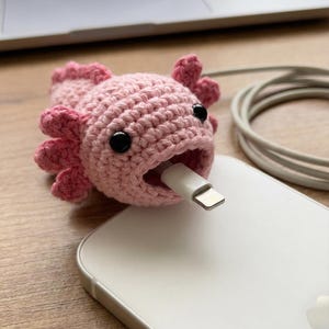 Może przedstawiać: Różowy, szydełkowy ochraniacz kabla axolotla z czarnymi oczami i białym kablem ładującym włożonym w jego pysk. Axolotl siedzi na białym smartfonie.