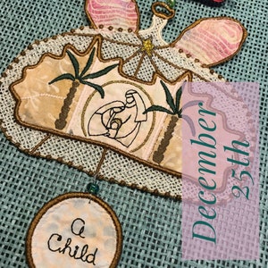 May include: A handmade Christmas ornament featuring a nativity scene with a star, palm trees, and the text "A child". The ornament is decorated with brown, gold, and pink embroidery and is hanging from a green and brown burlap background. The text "December 25th" is visible in the background.