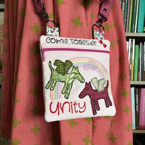 Könnte beinhalten: Eine rosa Stofftasche mit Reißverschluss und einem Regenbogen-Design. Die Tasche ist mit den Worten "Come Together" und "Unity" bestickt. Die Tasche ist an einem rosa und schwarzen Riemen befestigt.