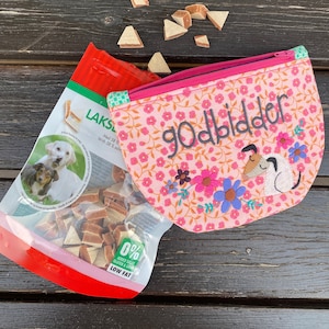 May include: A pink fabric pouch with a zipper closure and a floral pattern. The pouch has the word "godbidder" embroidered on it in black thread. The pouch is sitting on a wooden surface with a bag of dog treats in the background. The bag of treats is red and green and has a picture of a dog on it. The bag also has the text "LAKSL" and "0% ADDED SUGAR GLUTEN & GRAIN LOW FAT" on it.