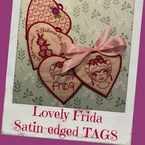 May include: Heart-shaped tags with satin edges, featuring embroidered designs. One tag displays a Frida Kahlo portrait, others have floral and text details. A pink ribbon adorns the tags. The text "Lovely Frida Satin-edged TAGS" is at the bottom.