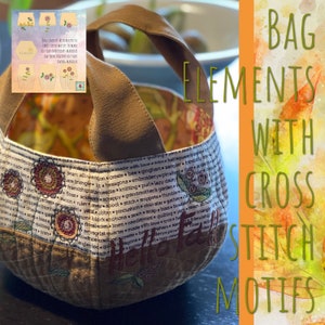 Pode incluir: Uma bolsa de tecido castanho com um design de ponto cruz que apresenta motivos florais e o texto "Hello Fall". A bolsa tem uma alça de lona castanha e é feita de tecido patchwork.